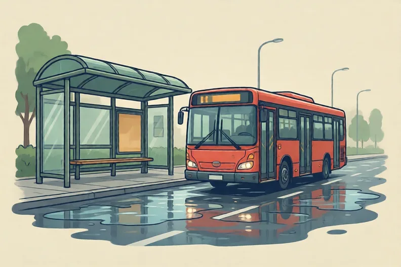 A bus stop on a wet street with a red bus pulling in